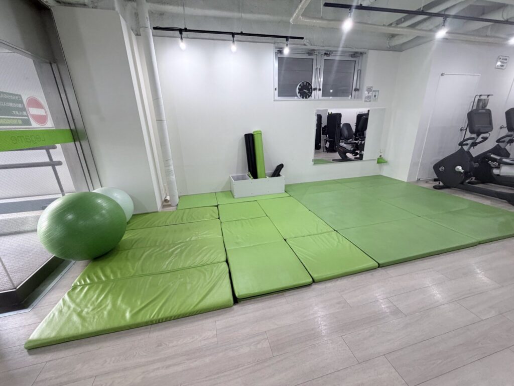 緑のマットが敷かれたストレッチコーナーと、奥に並ぶマシン。人はいない