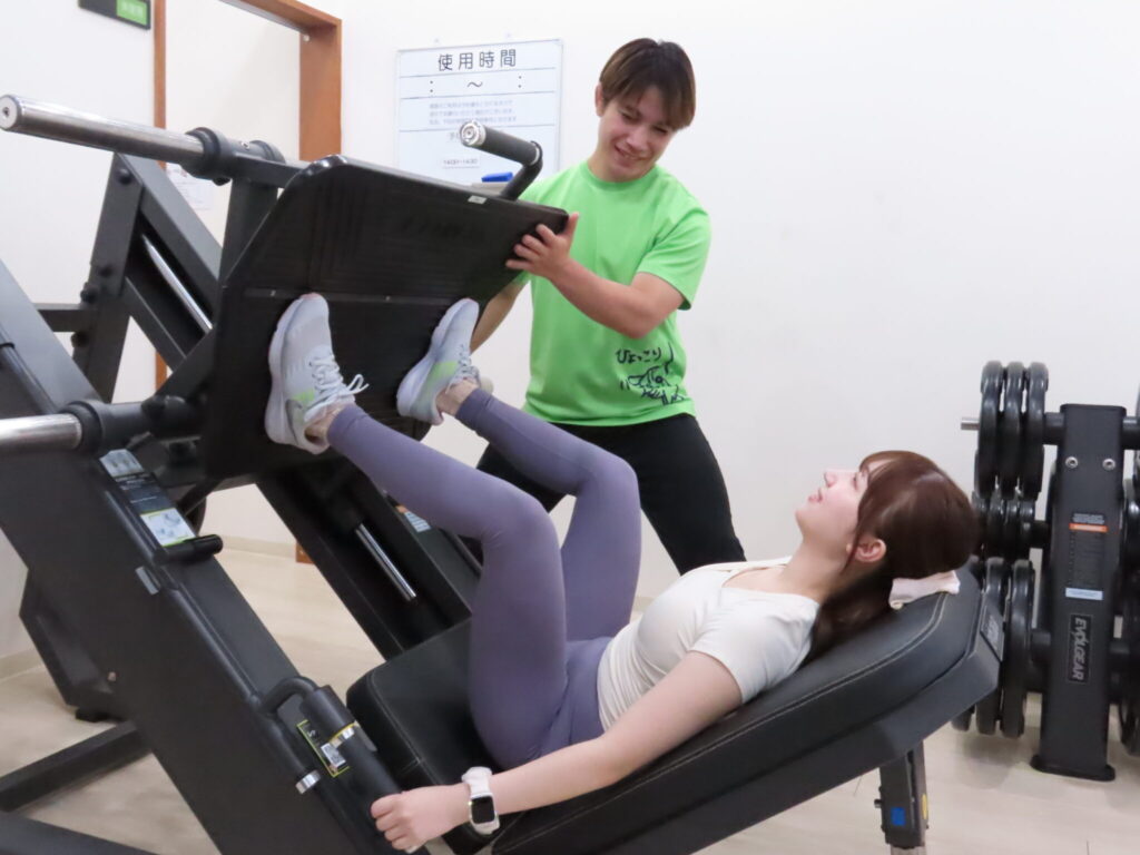 トレーナーがレッグプレスのプレートを支えながら、仰向けで脚を押す女性の動きを見ている