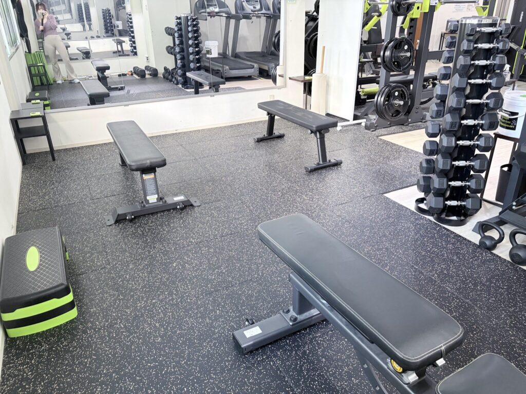 黒いラバーマットの上にフラットベンチが並び、右側にダンベルラック、奥にマシンとトレッドミルが見えるフリーウェイトエリア